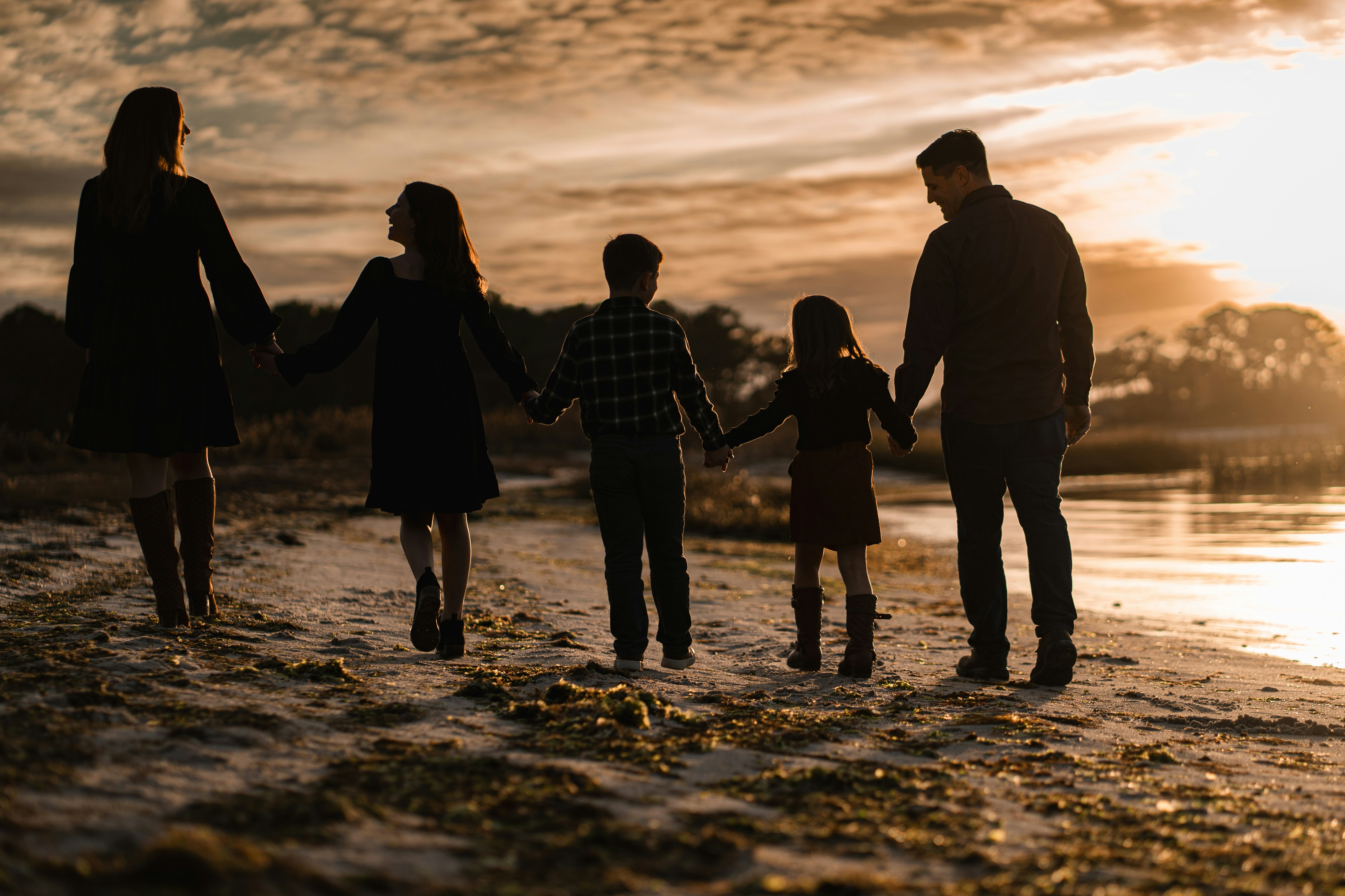 A family of five holding hands, backlit on a beach at sunset.